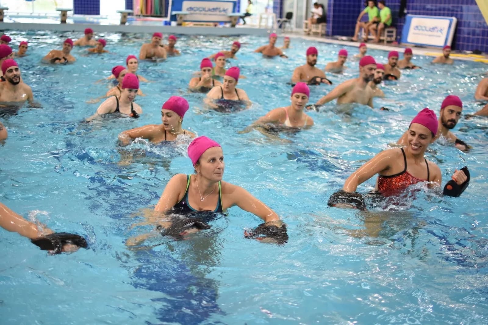 Aula de aquafitness com dezenas de participantes de touca cor-de-rosa dentro de água