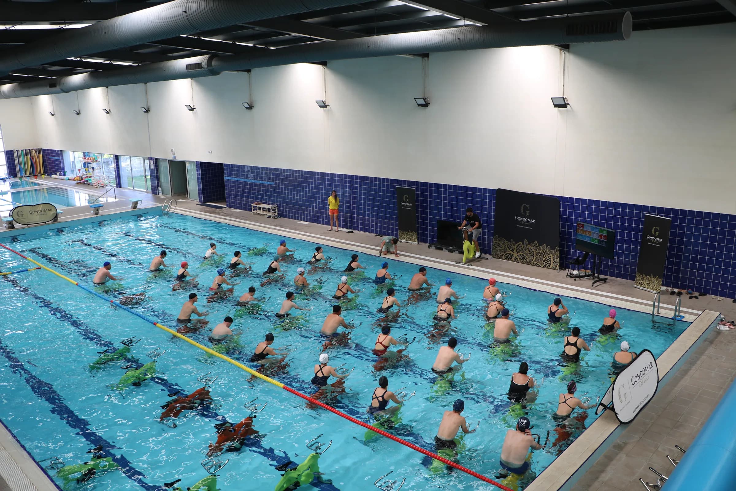 Vista de cima de uma piscina coberta durante uma aula de aquabike, com dezenas de participantes na água e formadores na borda
