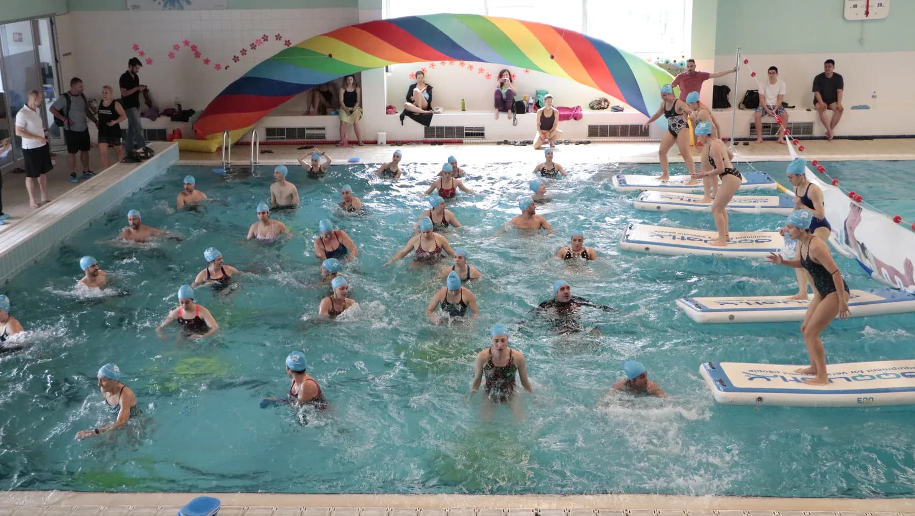 Sessão numa piscina com participantes de touca azul dentro de água e pranchas flutuantes junto à borda